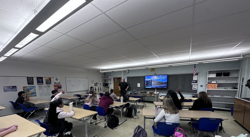 Classroom full of students raising their hands, with SRO Deputy Clark at the front of the classroom answering questions