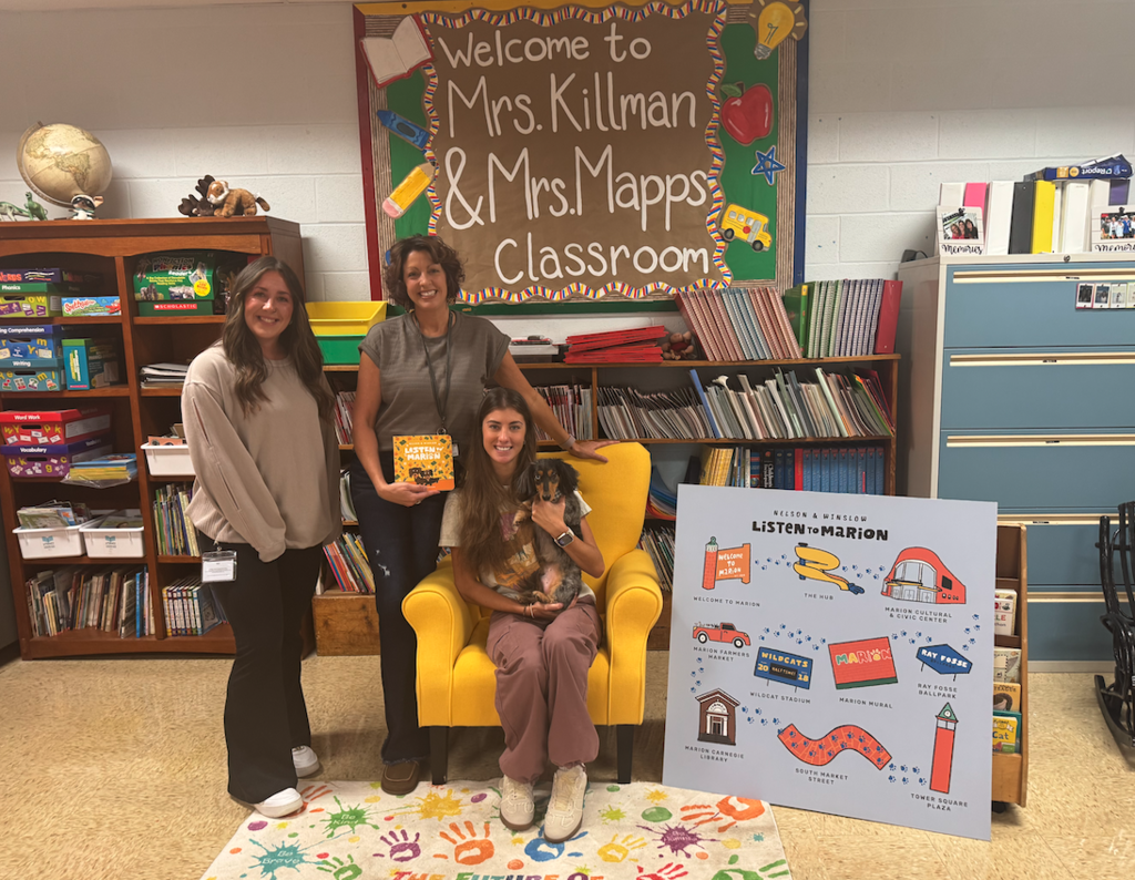 3 women in a classroom. 2 are standing and 1 is sitting in a yellow chair holding a dog