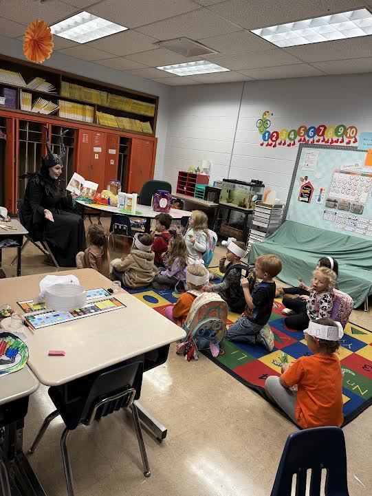 kindergarten class sitting on the floor listening to a mystery reader dressed as Maleficent