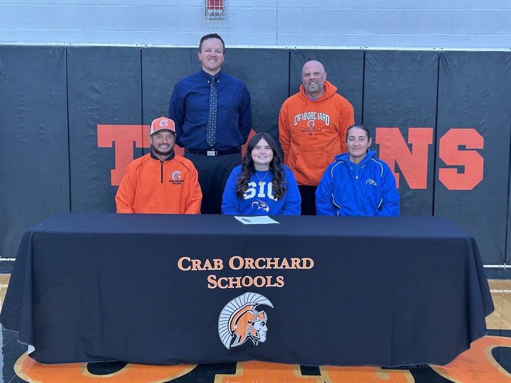 Picture of player signing her scholarship with blue sic shirt. Coaches next her have on orange crab shirt and sic shirt. Coaches behind have orange shirt 