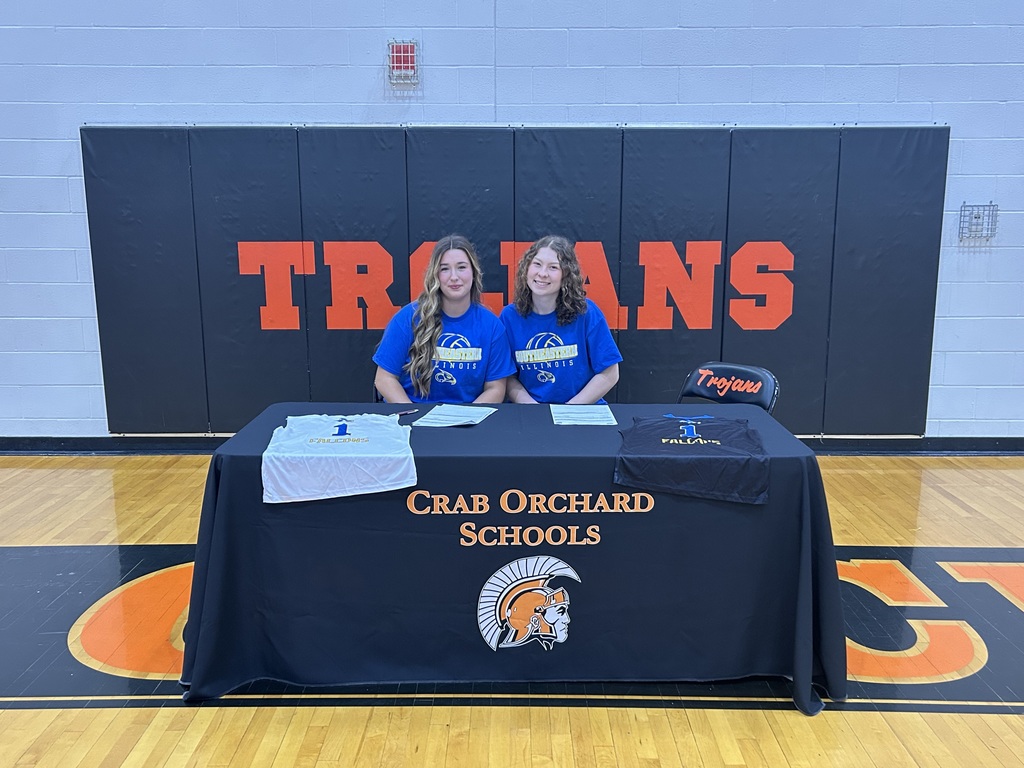 Two softball players in blue shirts sitting behind black table 