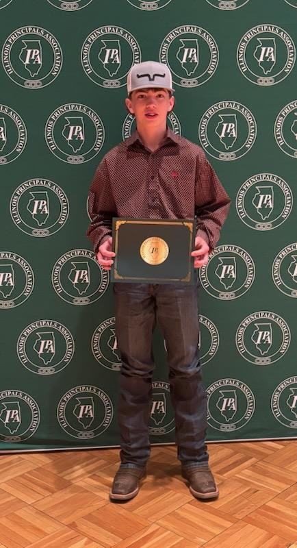 junior high male student holding a certificate standing in front of a green IPA sign