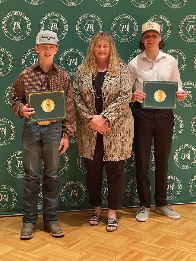 two males wearing hats and holding certificates standing with an adult female with long blonde hair in front of a green IPA sign