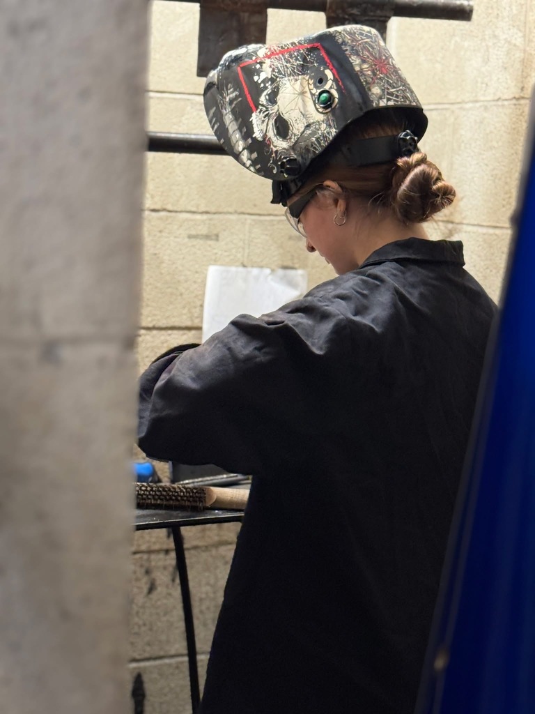 high school female wearing a welding hat, long dark jacket with her hair pulled back in a bun, standing in a concrete booth.
