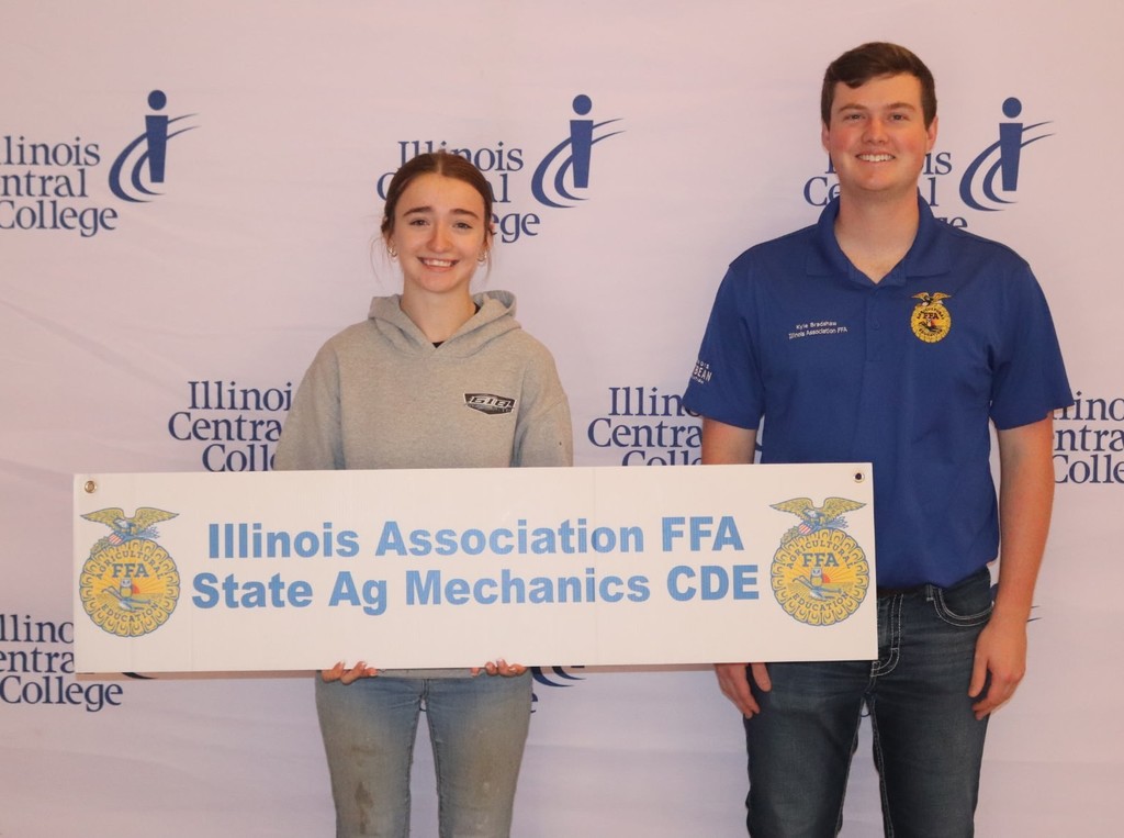young woman with dark hair pulled back wearing dirty jeans and a gray hoodie holding a sign with 2 FFA insignias and "Illinois Association FFA State Ag Mechanics CDE" standing beside a man  with dark hair wearing  a blue  FFA polo and jeans