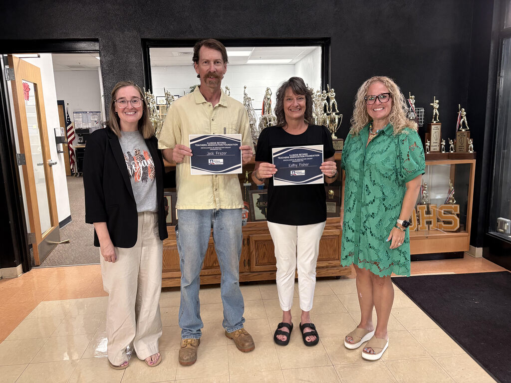 Four adults (one male and three females) standing in front of a trophy cabinet with two holding grant certificates