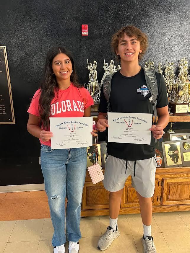 Two high school students, on female and one male, standing in front of a trophy case holding certificates.