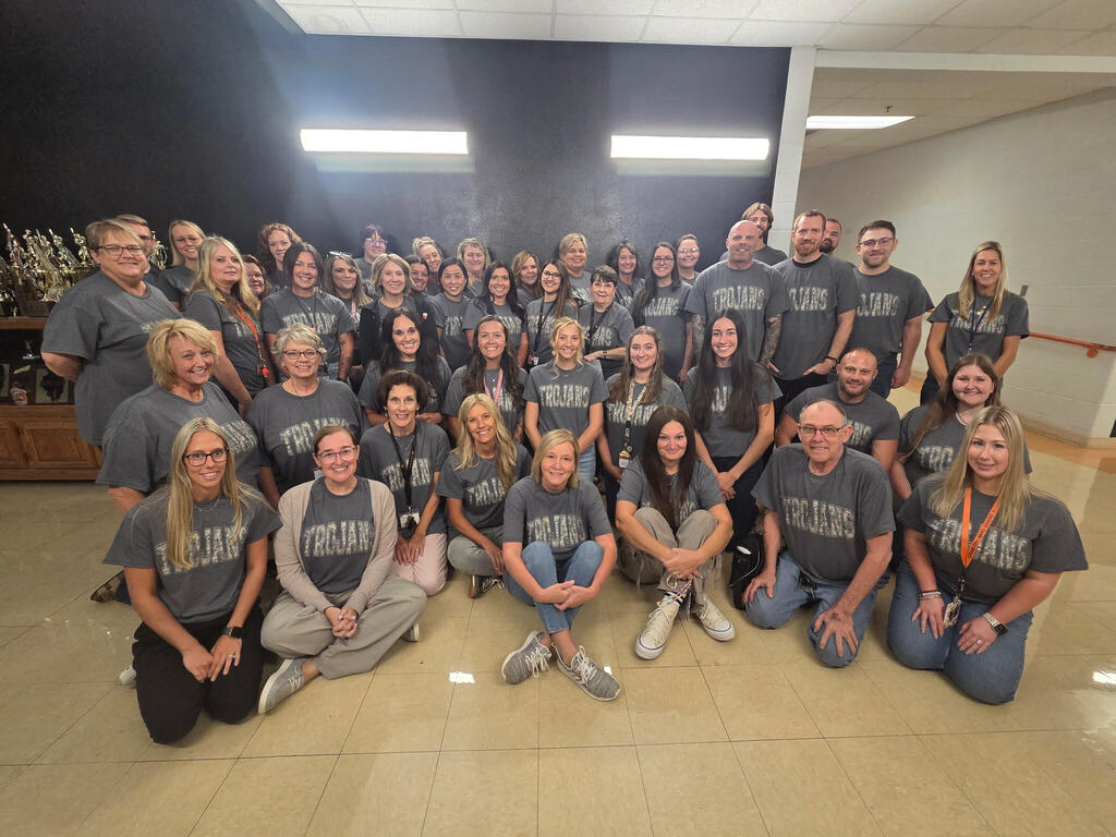 Group of teachers and staff sitting in a hallway with gray t-shirts posing for a group photo
