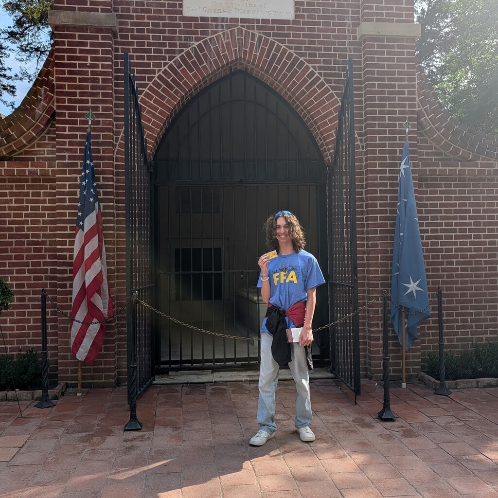 Young man in a light blue FFA shirt and jeans standing in front of George Washington's grave