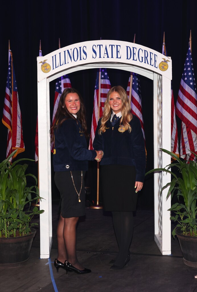 2 young ladies in official FFA attire under an Illinois State Degree arch in front of a row of US flags
