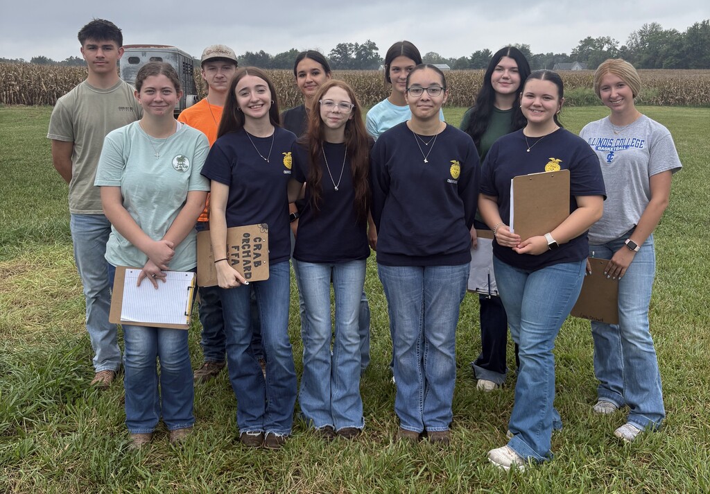 9 female and 2 male high school students wearing jeans and t-shirts standing in front of a field of corn and horse trailer