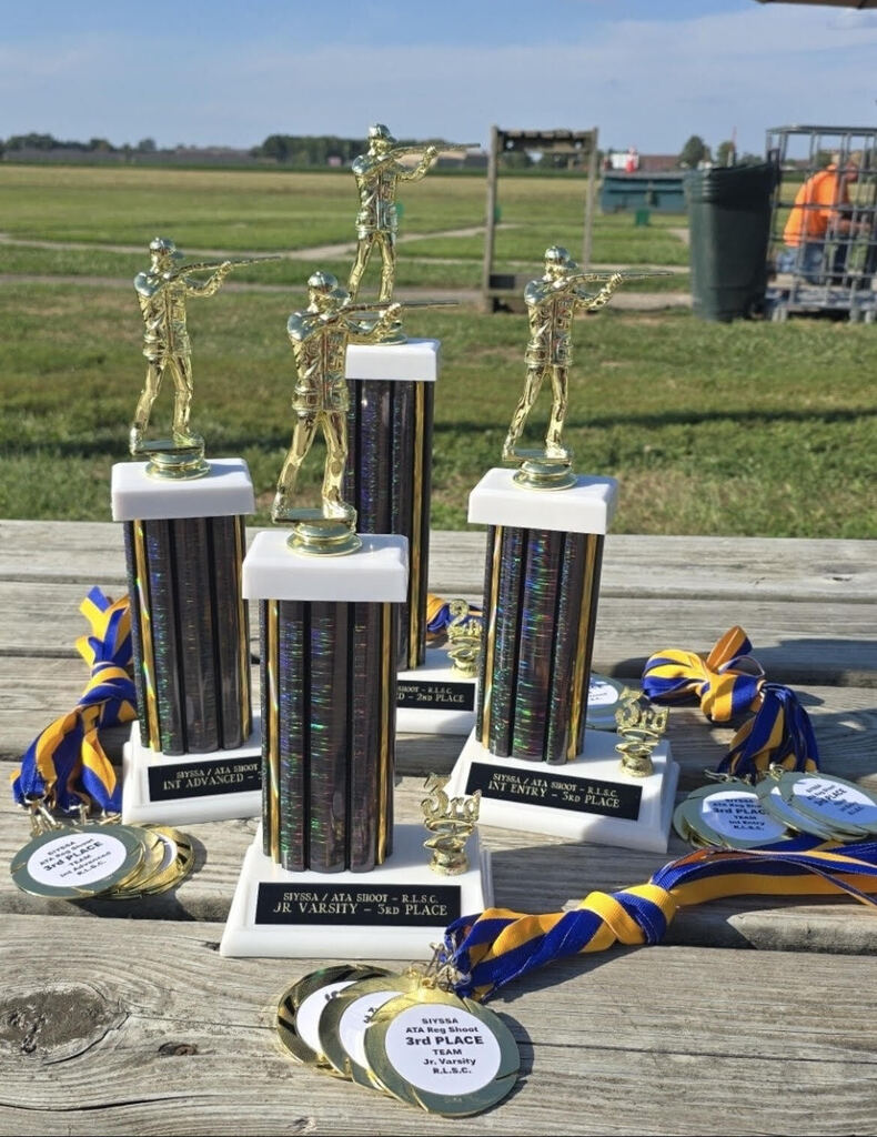 4 trophies with medals sitting on a wooden bench with grass in the background