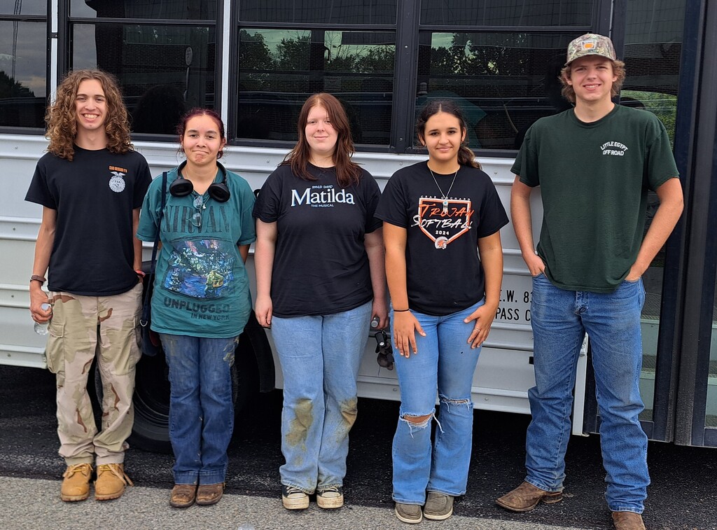 3 young ladies and 2 young men in dirty jeans and t-shirts standing in front of a white bus