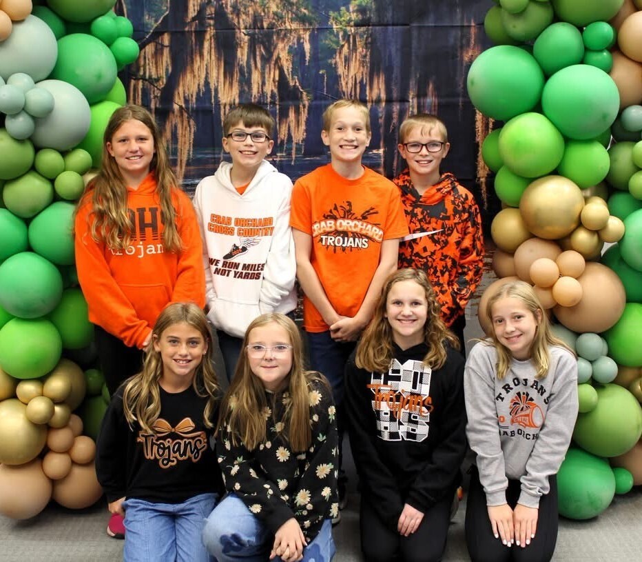 5th & 6th grade students standing & kneeling in front of green & gold balloons and a swamp backdrop
