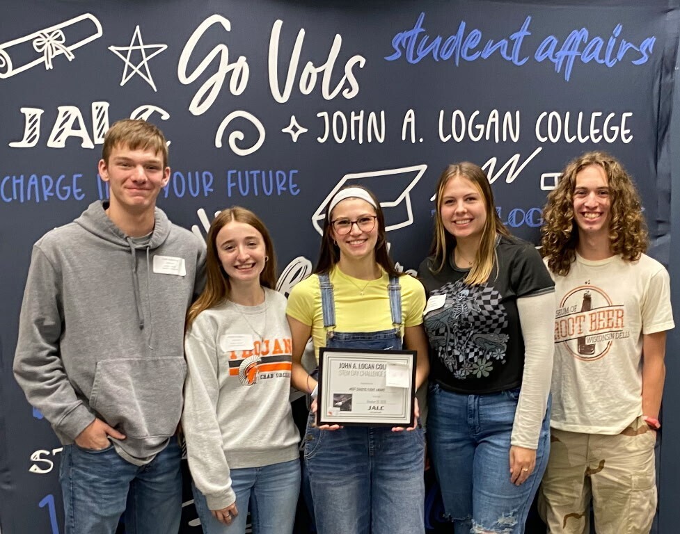 3 young ladies and 2 young men with a certificate in front of a John A Logan College banner
