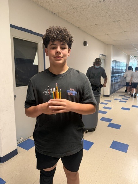 A boy with curly hair holds yellow pencils in a hallway. Behind him, a person stands near a locker.