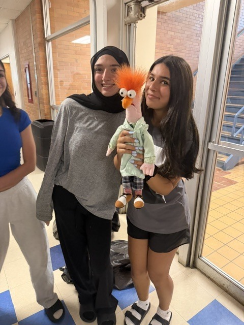 Two girls stand in a hallway, one holding a Muppets character puppet, another woman in a hijab.