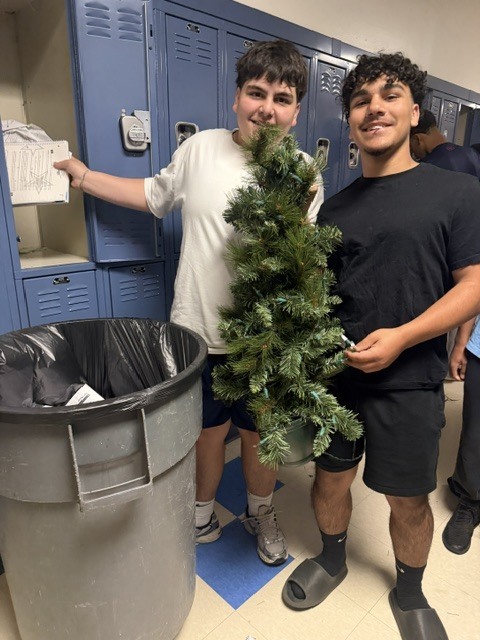 Two individuals, one holding a small artificial Christmas tree, stand next to a trash bin. Blue lockers are behind them.