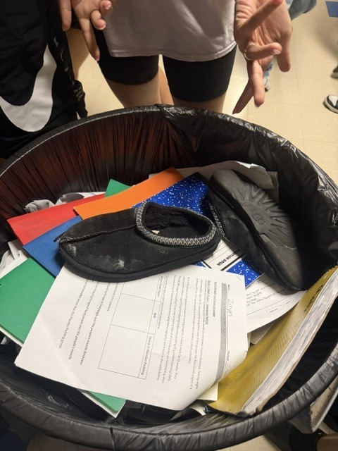 A black trash can filled with various items, including a black sandal, colorful papers, and other objects.