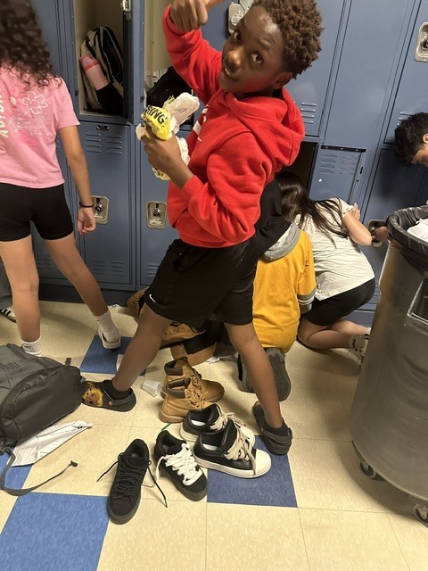 A child stands in a school hallway with a toy, surrounded by bags, shoes, and a trash bin.