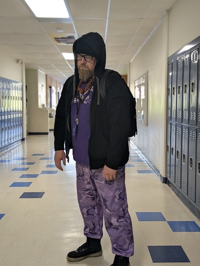 Man in a hoodie, purple shirt, purple pants, and black shoes with a backpack.