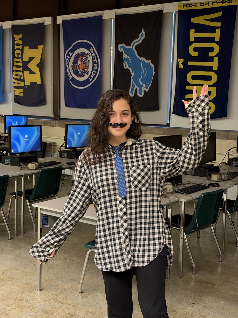 Person in black and white checkered shirt, blue tie, and black pants standing in a classroom.