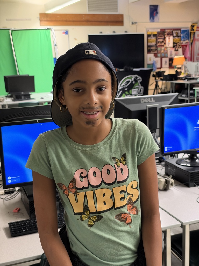 A young girl wearing a green T-shirt stands in a computer lab, smiling at the camera.