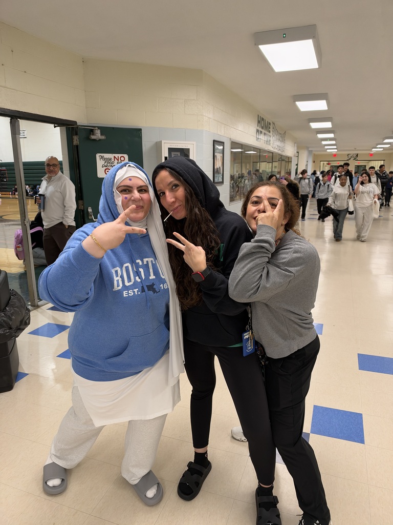 Three girls stand in a hallway, smiling, with one making a peace sign. Behind them, people walk.