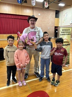 Picture of older man holding a dinosaur puppet with kids around him. 