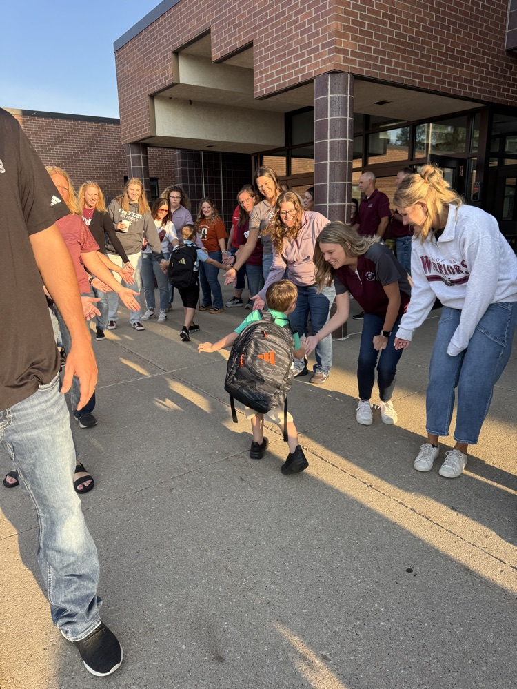 Members of Crofton Community Schools are smiling in front of the high school to greet and high five the students