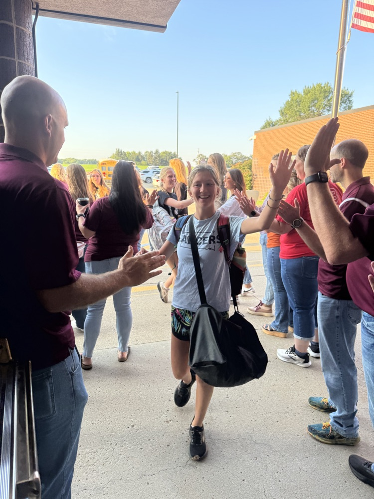 Members of Crofton Community Schools are smiling in front of the high school to greet and high five the students