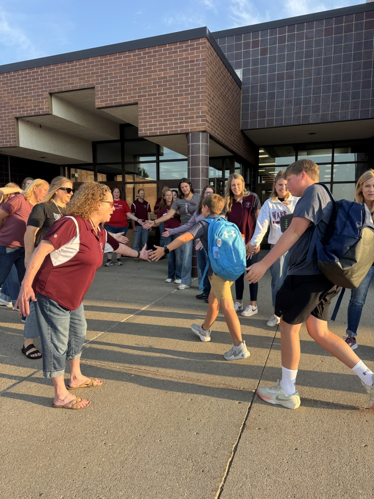 Members of Crofton Community Schools are smiling in front of the high school to greet and high five the students