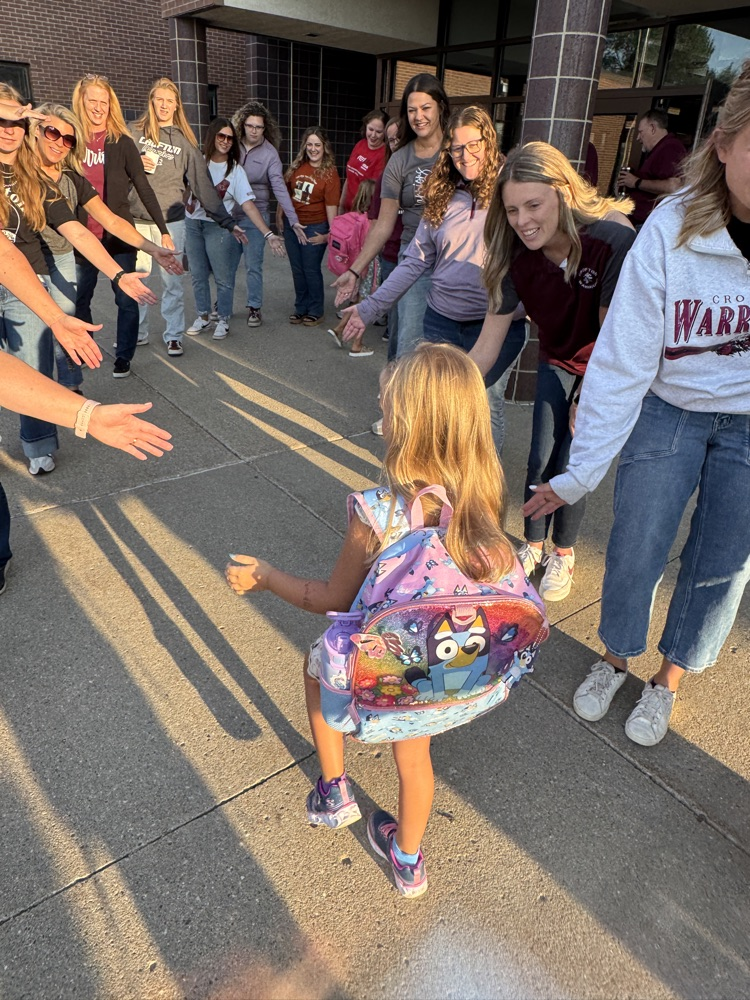 Members of Crofton Community Schools are smiling in front of the high school to greet and high five the students
