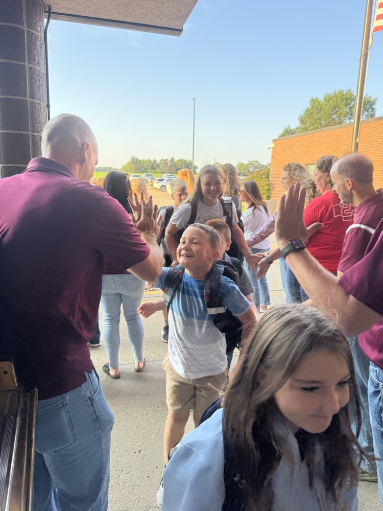 Members of Crofton Community Schools are smiling in front of the high school to greet and high five the students