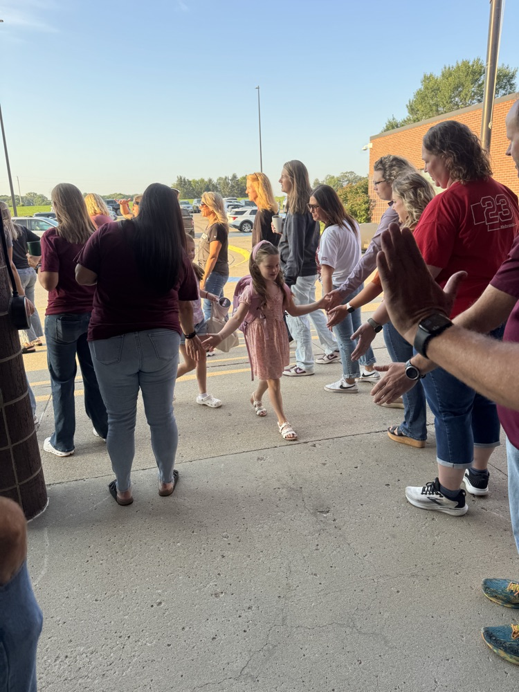 Members of Crofton Community Schools are smiling in front of the high school to greet and high five the students