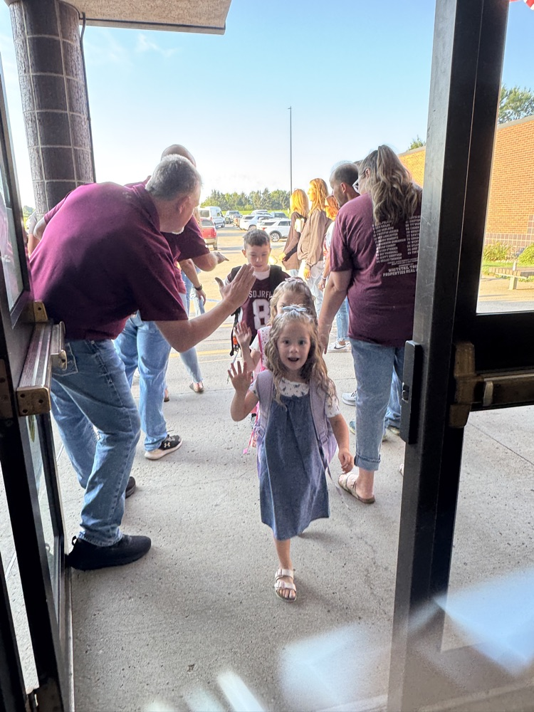 Members of Crofton Community Schools are smiling in front of the high school to greet and high five the students