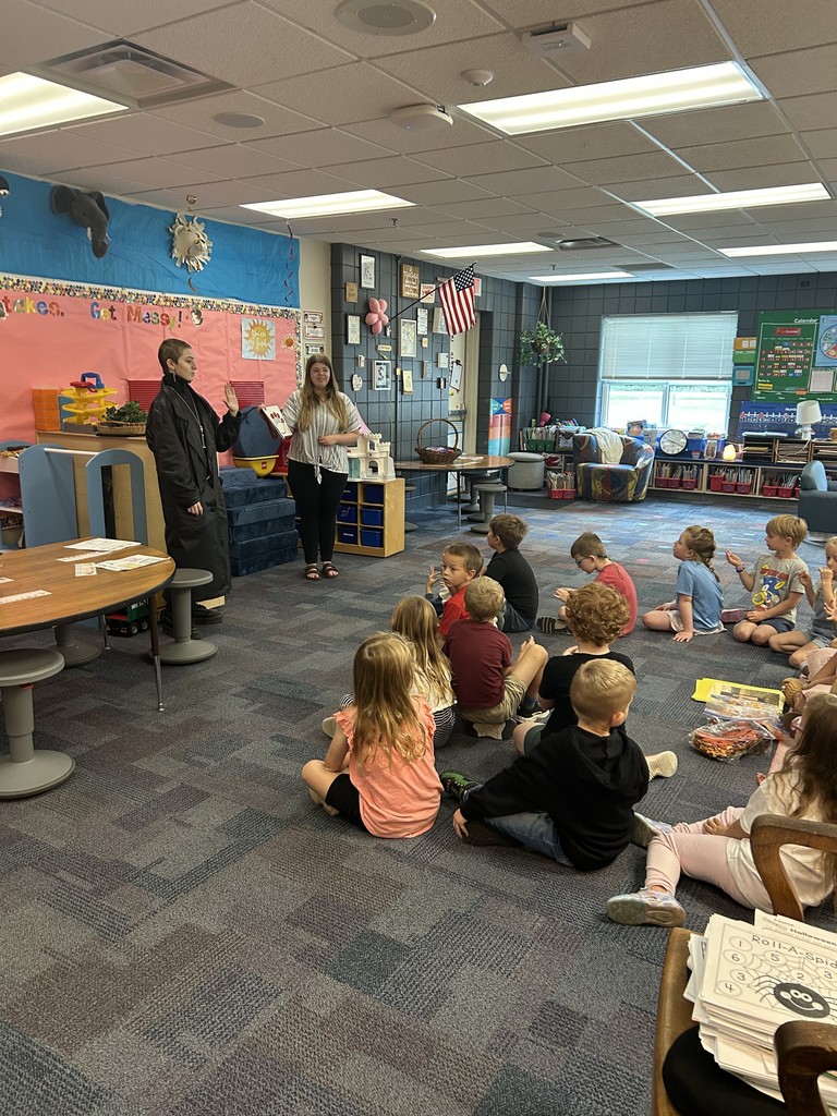 two high school students standing in front of younger students who are sitting on the floor