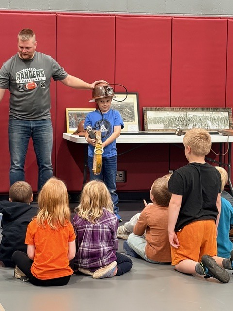 One of the 6th grade teachers standing next to a young student who is wearing an old miners hat with a light on it in front of a group of kids watching