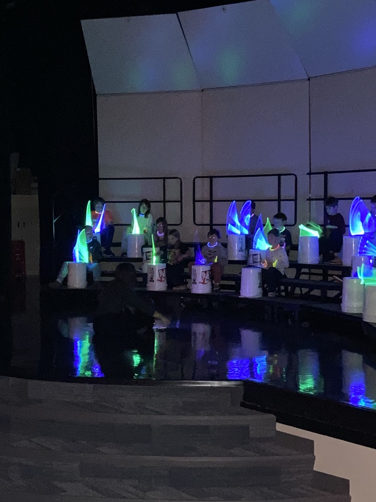3rd graders playing drums using glow in the dark blue and green drumsticks at their expo