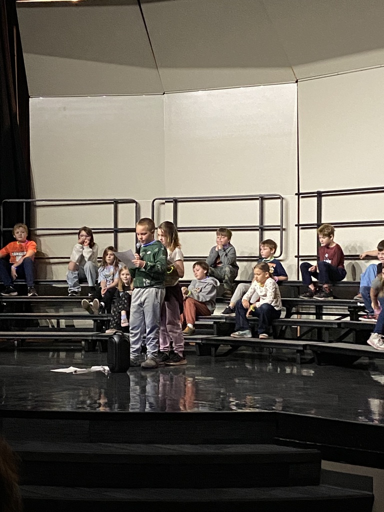 third grade student standing on stage in the performance area reading from a sheet of paper at their expo