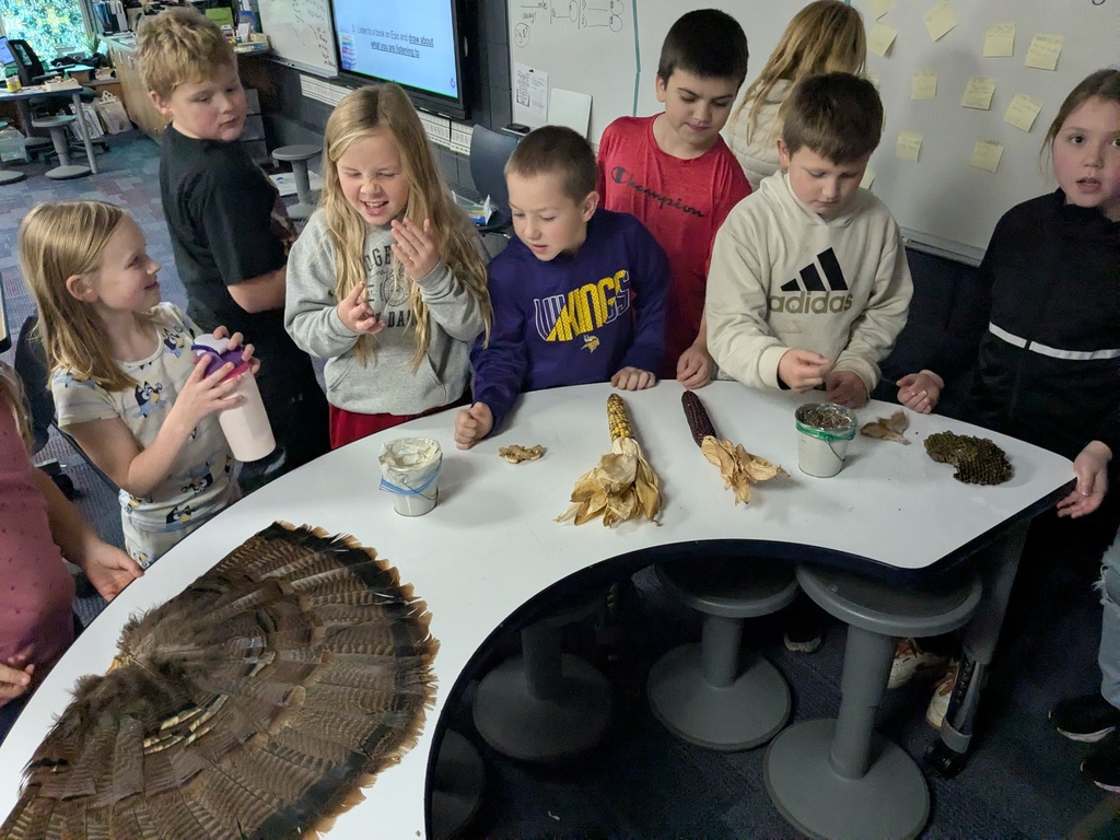 3-4th graders standing around a table with turkey feather,s corn stalks and other items that remind them of Thanksgiving