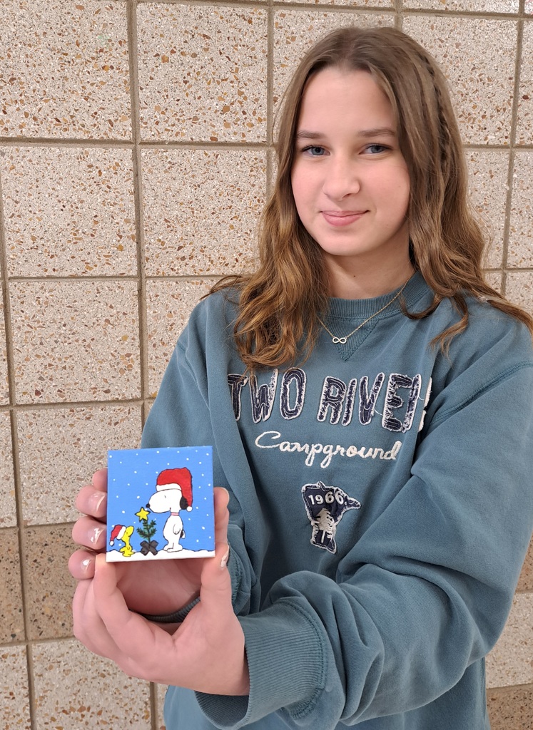 female student holding a mini acrylic painting of snoopy with a santa hat and his yellow bird friend with a santa hat on next to a tiny little Christmas tree