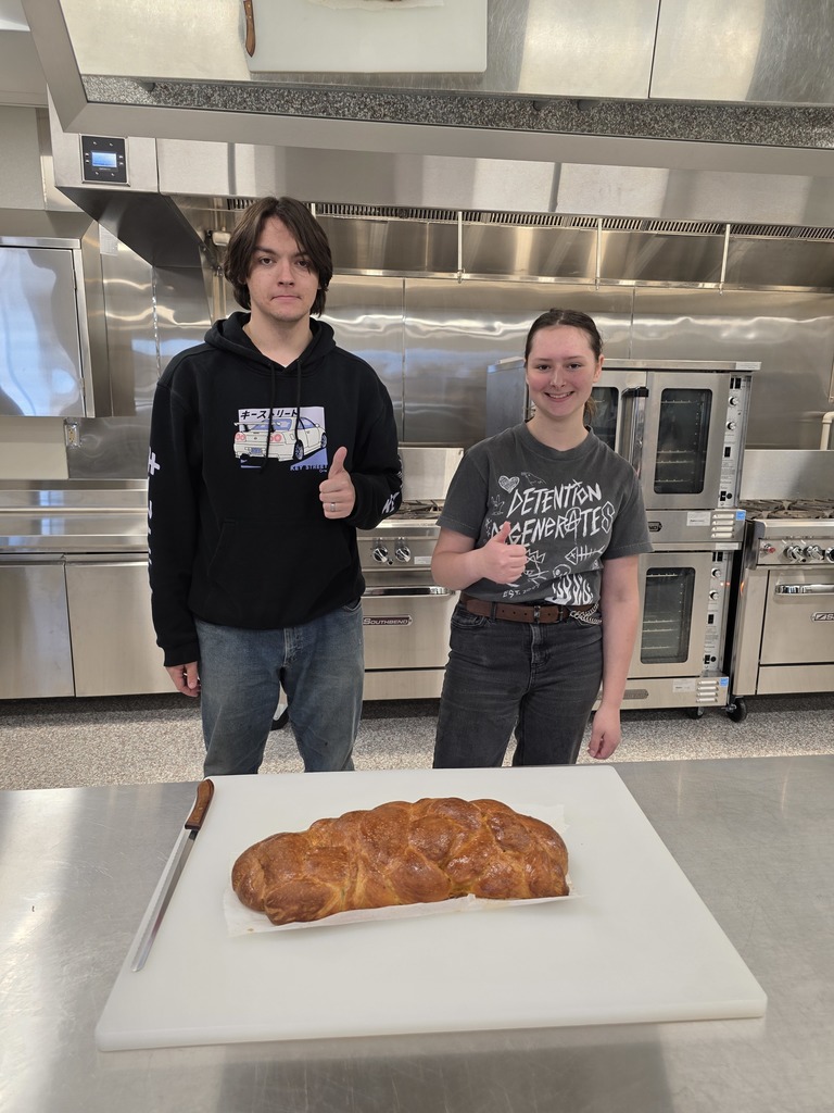 one male and one female student who is giving thumbs up to the loaf of Challah bread on the cutting board in front of them