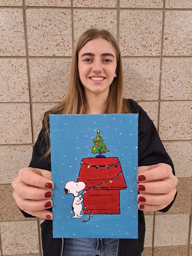 Female 10th grade student holding a painting of Snoopy putting colored christmas lights on his red dog house with a Christmas tree on top