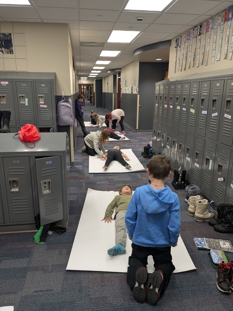 a hallway lined with students laying on big white sheets of paper as their classmates trace them