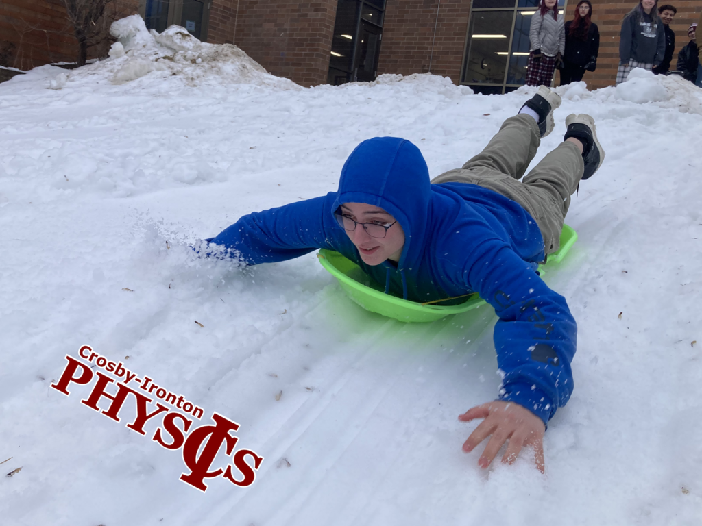 male student in a blue coat sledding on a green sled on his stomach in physics class