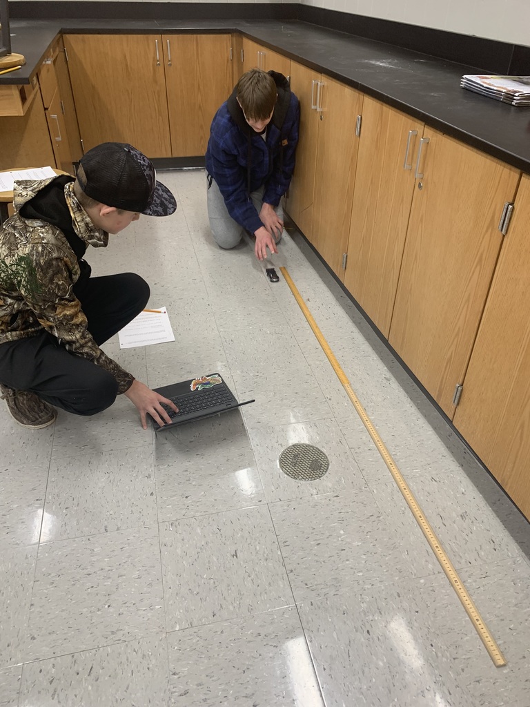 two male students sitting on the floor, one working on the computer the other winding up a toy car and letting it go so they can measure the speed