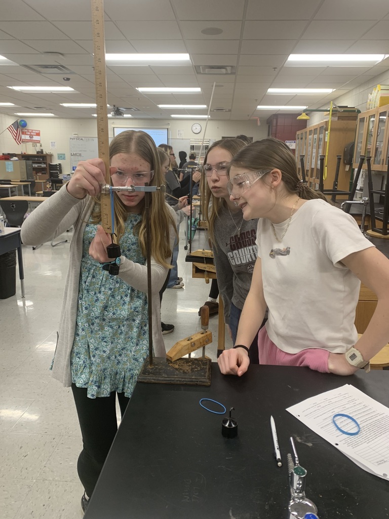 three females students taking measurements of a stretched out rubberband with weights on it