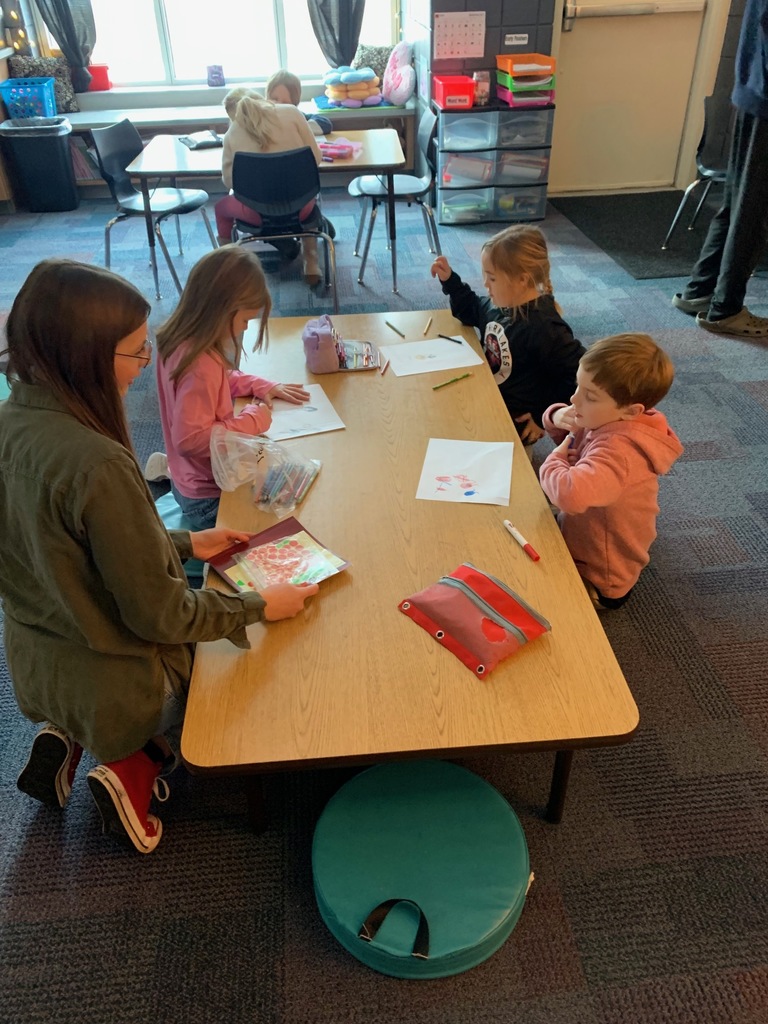 Hs female student kneeling at a table with younger students helping them with a lesson