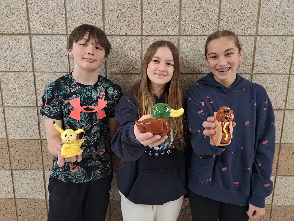 three 7th grade students holding their clay projects- a Pokeman, duck, weiner dog in a bun